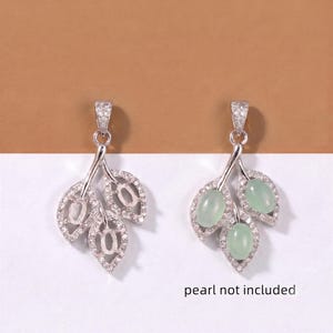 Puede incluir: Dos colgantes plateados en forma de hoja con piedras pavé transparentes. Un colgante tiene engastes vacíos, mientras que el otro contiene tres cabujones ovalados de color verde pálido. El texto "pearl not included" aparece en negro abajo.
