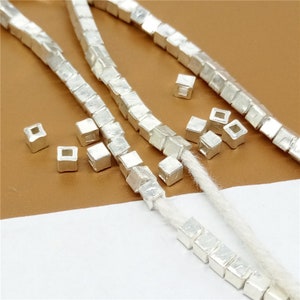 Può includere: Perle quadrate d'argento con un foro al centro. Le perle sono infilate su un filo bianco.