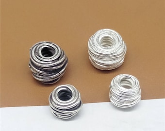 Może przedstawiać: Cztery srebrne metalowe koraliki o okrągłym, spiralnym wzorze. Koraliki są różnych rozmiarów i ułożone są w rzędzie na brązowej powierzchni.