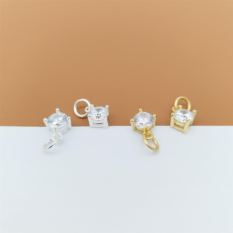 以下が含まれることがあります： 銀色の金属製チャーム2個と金色の金属製チャーム2個。それぞれ、4本爪の台座にセットされた透明なラウンドカットのキュービックジルコニアが1粒あしらわれており、上部には小さな丸カンが付いています。