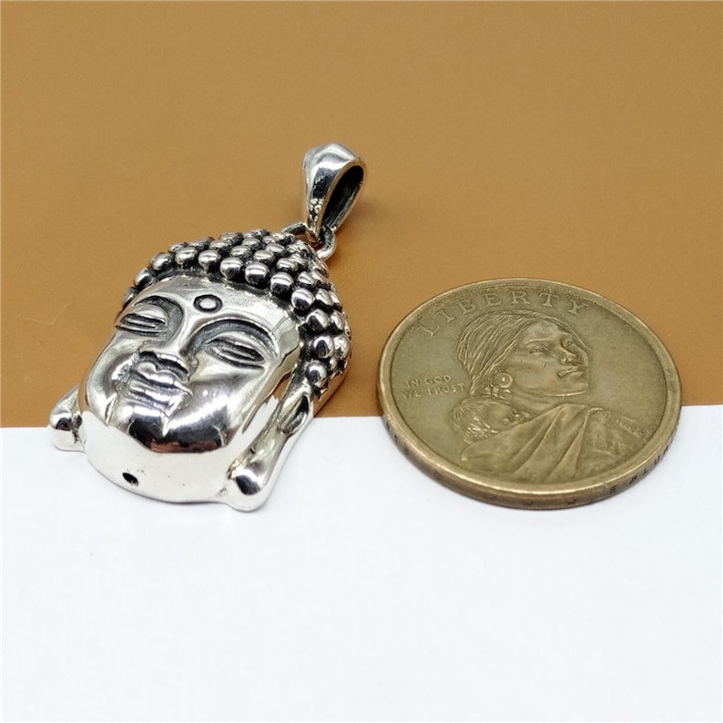 May include: Silver pendant featuring a detailed Buddha head with closed eyes. The pendant is shown next to a US dollar coin for size comparison.