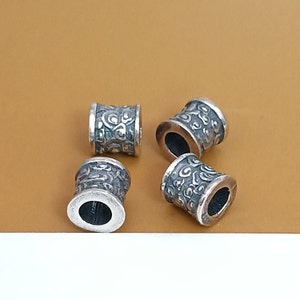Può includere: Quattro perline in metallo color argento con un designo testurizzato e decorato. Le perline sono cilindriche con una parte superiore e inferiore leggermente più ampia.