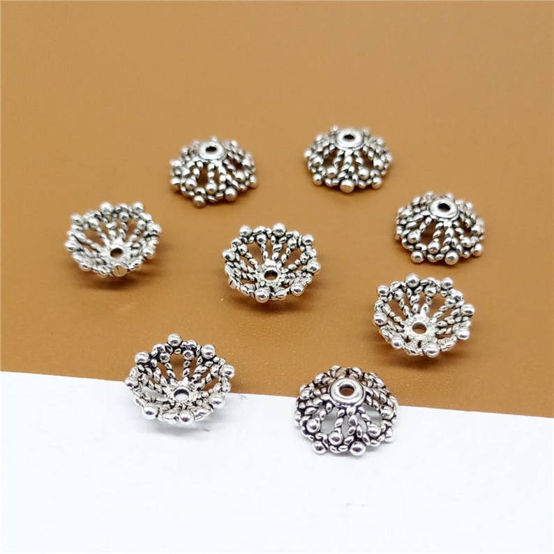 May include: Ten silver-toned metal bead caps with a decorative, circular, openwork design. The caps are arranged on a brown surface.