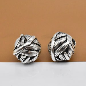 Puede incluir: Dos cuentas de metal plateadas con un diseño tejido detallado. Las cuentas tienen un patrón texturizado e intrincado que se asemeja a una bola de hilo.