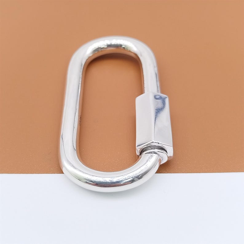 May include: A shiny, silver-colored oval carabiner with a rectangular locking mechanism. The carabiner is made of a smooth, reflective metal and is designed for securing items. The background is a light brown and white.