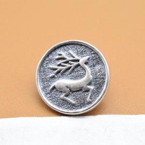May include: A silver-toned button with a raised design of a deer with antlers. The button has a textured surface.