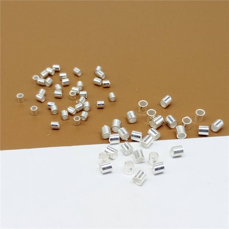May include: A large quantity of silver-colored metal jump rings. The jump rings are small and cylindrical in shape. They are scattered on a brown surface and a white surface.