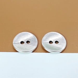 Può includere: Due bottoni rotondi color argento con due fori ciascuno. I bottoni sono rotondi e hanno un bordo leggermente rialzato.