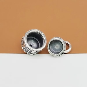 Puede incluir: Un charm de plata con un dise&ntilde;o detallado y una peque&ntilde;a abertura. El charm est&aacute; en dos piezas, una superior y una inferior, que encajan entre s&iacute;. El charm est&aacute; dise&ntilde;ado para contener un peque&ntilde;o objeto, como una foto o un mensaje.