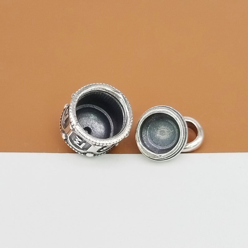 Puede incluir: Un charm de plata con un dise&ntilde;o detallado y una peque&ntilde;a abertura. El charm est&aacute; en dos piezas, una superior y una inferior, que encajan entre s&iacute;. El charm est&aacute; dise&ntilde;ado para contener un peque&ntilde;o objeto, como una foto o un mensaje.
