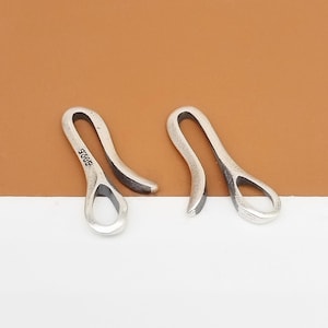 Könnte beinhalten: Zwei silberfarbene Metallhaken mit glattem, geschwungenem Design. Die Haken befinden sich auf einer braunen Oberfläche. Ein Haken trägt die Aufschrift "925S" an der Seite.