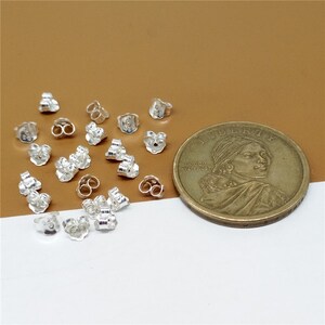 May include: A close-up of a pile of silver earring backs, next to a US dollar coin. The earring backs are small and have a simple design.