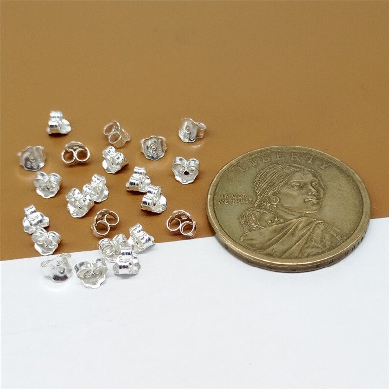 May include: A close-up of a pile of silver earring backs, next to a US dollar coin. The earring backs are small and have a simple design.