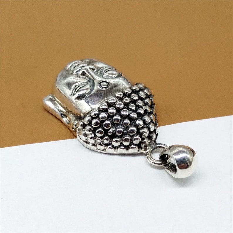 May include: Silver pendant featuring a detailed Buddha head with a textured design. The pendant has a loop for attaching to a chain.