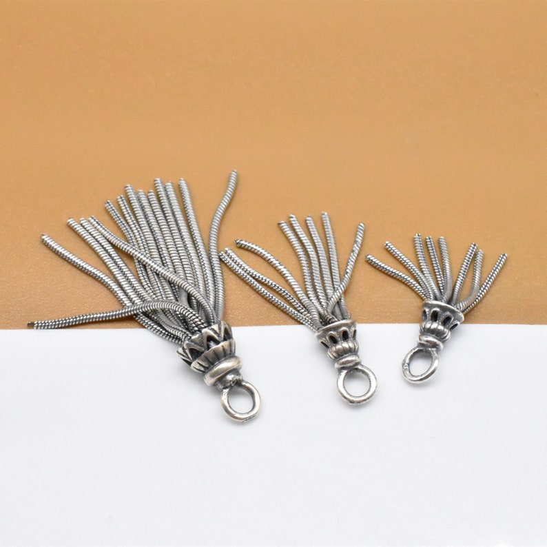May include: Three silver tassel charms of varying sizes with a decorative ring at the bottom. The tassels are made of thin, twisted wire.