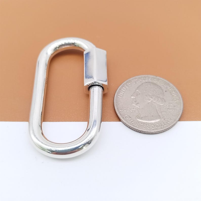 May include: A shiny, silver-colored oval carabiner clasp with a screw-lock mechanism. The clasp is next to a U.S. quarter coin for size comparison. The background is a mix of white and brown.