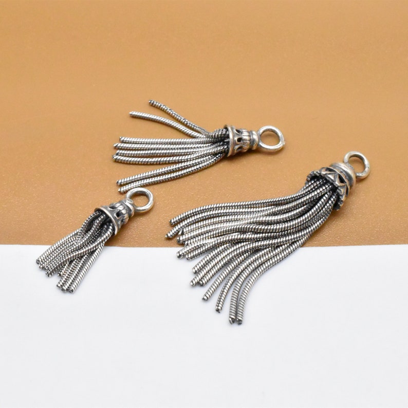 May include: Three silver tassel charms with a decorative top and a loop for attaching to a bracelet or necklace.