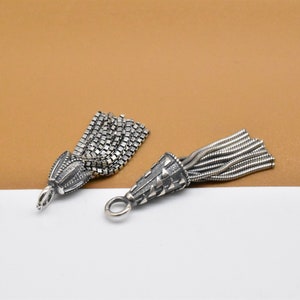 May include: Two silver tassel charms with different styles. One charm has a chain tassel and the other has a thin wire tassel. Both charms have a cone-shaped top with a decorative pattern.