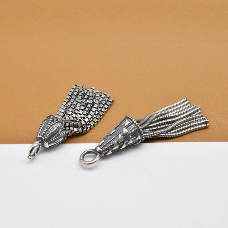 May include: Two silver tassel charms with different styles. One charm has a chain tassel and the other has a thin wire tassel. Both charms have a cone-shaped top with a decorative pattern.
