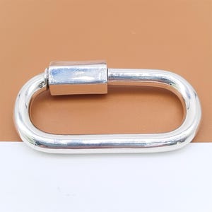May include: A shiny, silver-colored oval carabiner with a rectangular screw-lock mechanism. The carabiner is shown against a brown and white background, highlighting its polished metal finish. This hardware item is suitable for various crafting and jewelry projects.