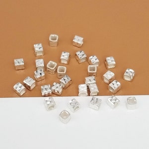 Könnte beinhalten: Ein Haufen silberner quadratischer Abstandshalterperlen mit einem rautenförmigen Muster. Die Perlen sind auf einer braunen und weißen Oberfläche verstreut.