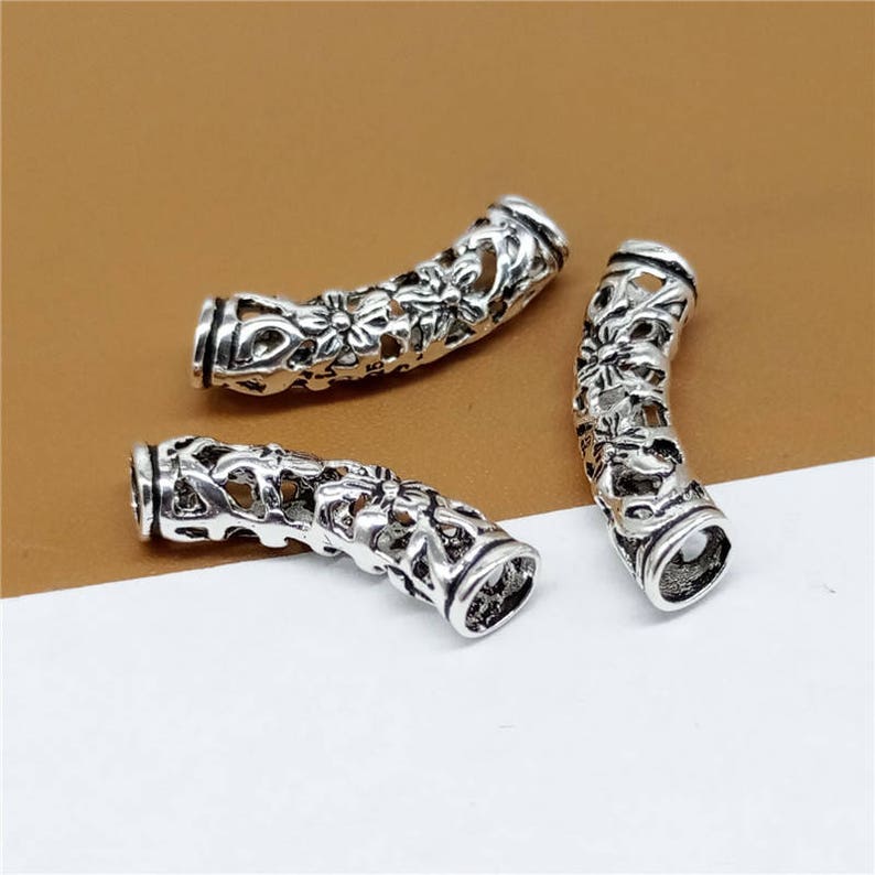 May include: Three silver-toned metal beads with intricate floral designs. The beads are shaped like a curved tube and have a hollow center.