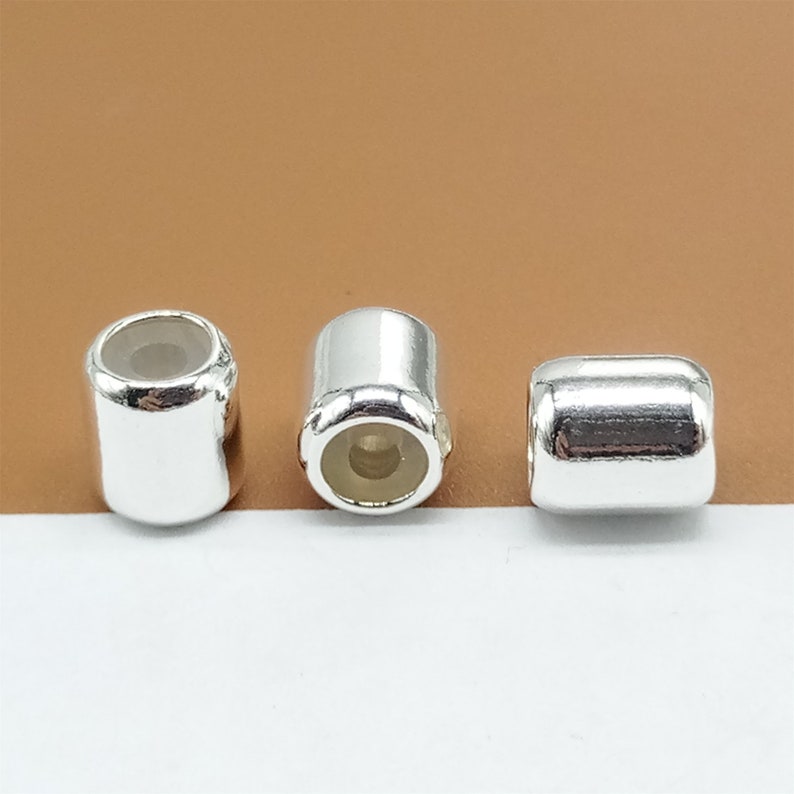 Op de afbeelding: Drie glanzende, zilverkleurige, cilindrische kralen met afgeronde randen. Elke kraal heeft een hol midden, en de uiteinden zijn open. De kralen worden gepresenteerd tegen een neutrale achtergrond, waardoor hun reflecterende oppervlakken worden benadrukt.