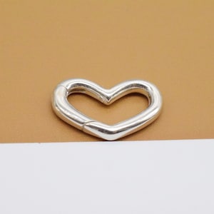 May include: A silver heart-shaped charm with a smooth, polished finish. The charm is open on one side, suggesting it can be used as a clasp or connector.