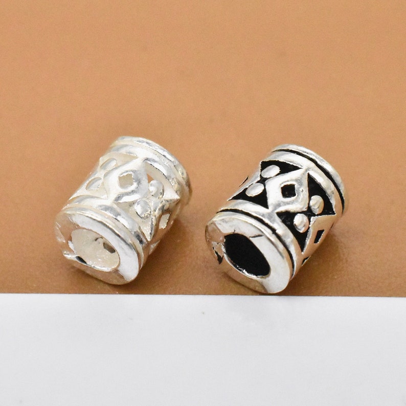 May include: Two silver-toned metal hair beads with intricate geometric designs. The beads are cylindrical and have a hollow center.