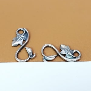 Puede incluir: Dos abalorios de metal de color plateado en forma de hoja con un bucle en la parte superior. Los abalorios tienen forma de vid con hojas.
