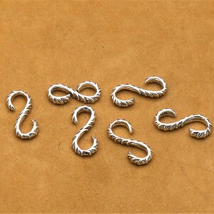 May include: Seven silver-toned metal hooks with a twisted rope design. The hooks are arranged in a semi-circle on a brown surface.