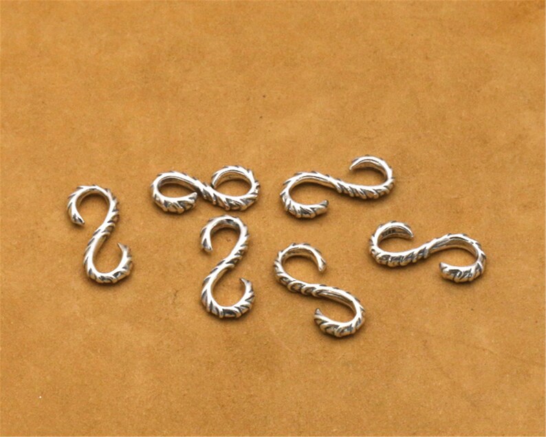 May include: Seven silver-toned metal hooks with a twisted rope design. The hooks are arranged in a semi-circle on a brown surface.