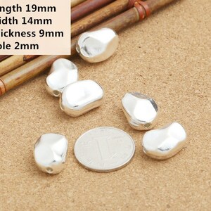 May include: Six silver-colored metal beads, each with a slightly irregular oval shape and a small hole for stringing. The beads are arranged on a light brown surface with a Chinese coin in the center.