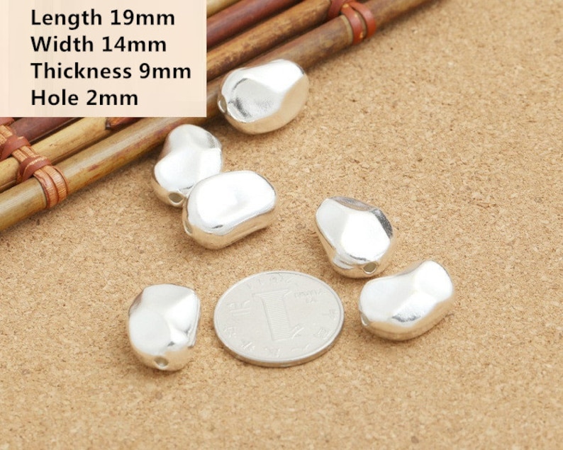 May include: Six silver-colored metal beads, each with a slightly irregular oval shape and a small hole for stringing. The beads are arranged on a light brown surface with a Chinese coin in the center.