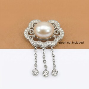 May include: A silver-toned metal pendant featuring a central circular setting for a pearl, surrounded by an intricate filigree frame encrusted with small clear stones. Three delicate silver chains hang from the base, each ending in a single clear stone. The text "pearl not included" is visible.
