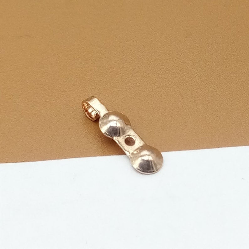 May include: A gold-toned metal pendant with two rounded ends and a loop at the top. The pendant has a small hole in the center.