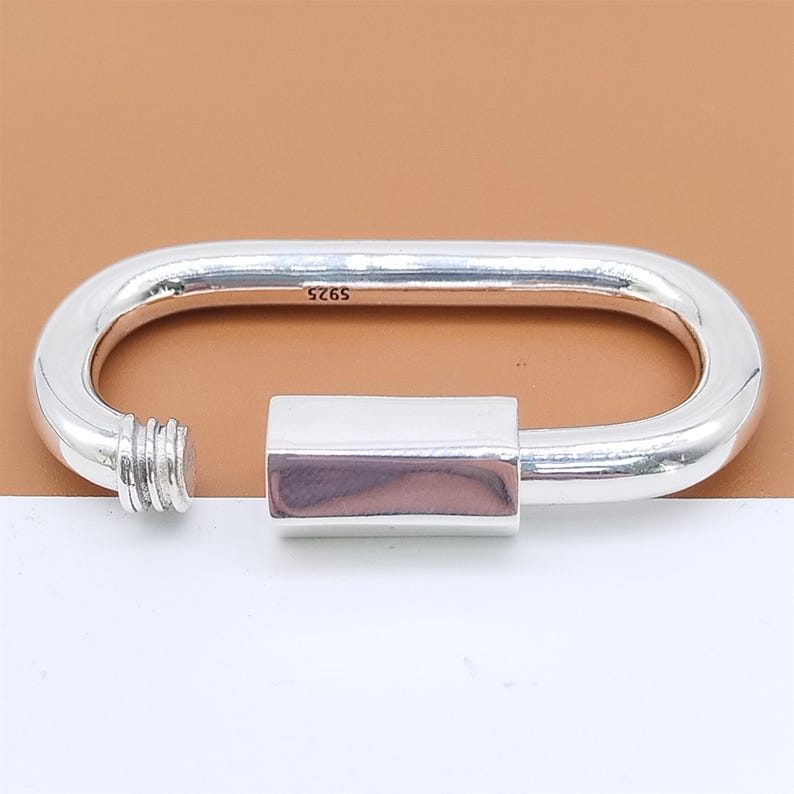 May include: A silver-colored, oval-shaped clasp with a screw-on end and a rectangular closure. The clasp is made of a shiny metal and has the number "925" stamped on it.