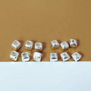 May include: A collection of silver-toned square-shaped beads with a hole in the centre. The beads are arranged in two rows on a gold surface.