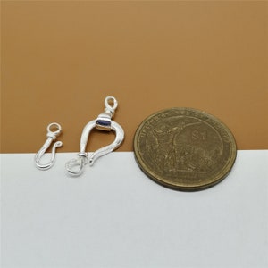 May include: Two silver-colored jewelry clasps, one small and one larger, are displayed next to a US one-dollar coin. The clasps have a hook and eye design, with a curved shape. The background is a light brown and white.