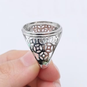 Puede incluir: Un engaste de anillo plateado con un diseño calado. El anillo tiene forma cónica con una parte superior circular y un patrón decorativo de formas geométricas. El anillo se sostiene sobre un fondo blanco.