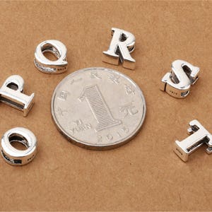 May include: Silver charm beads in the shape of the letters P, Q, R, S, T, and I. The beads are on a brown surface.