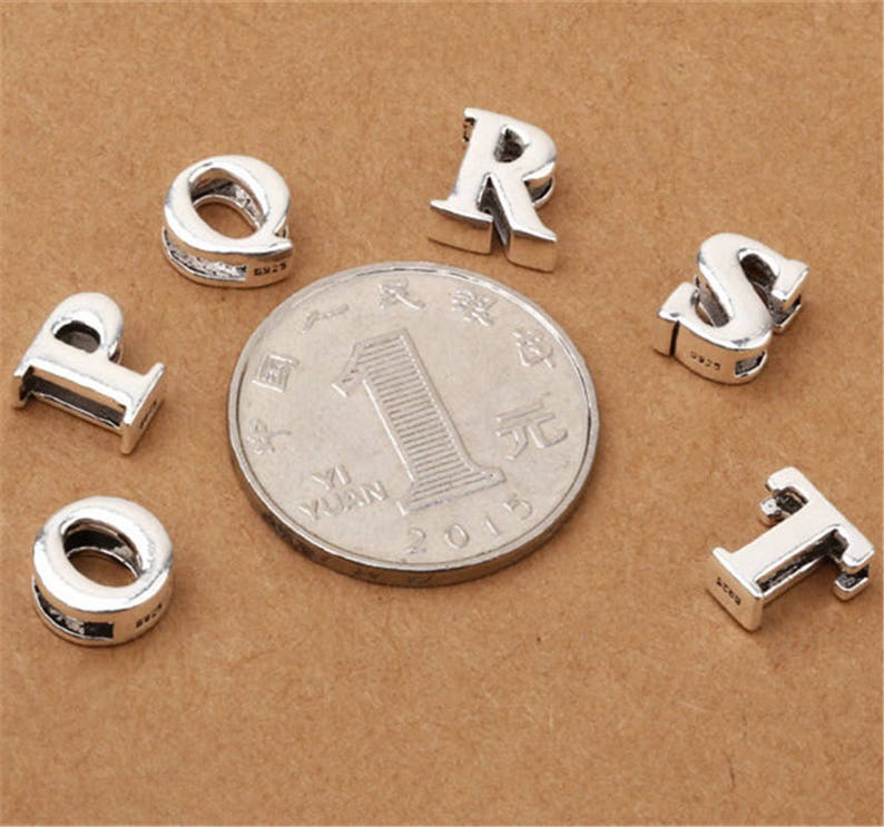 May include: Silver charm beads in the shape of the letters P, Q, R, S, T, and I. The beads are on a brown surface.