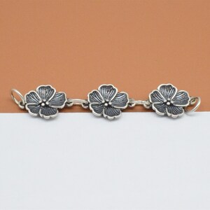 May include: A silver chain with three flower-shaped charms. The flowers are detailed and have a black oxidized finish.