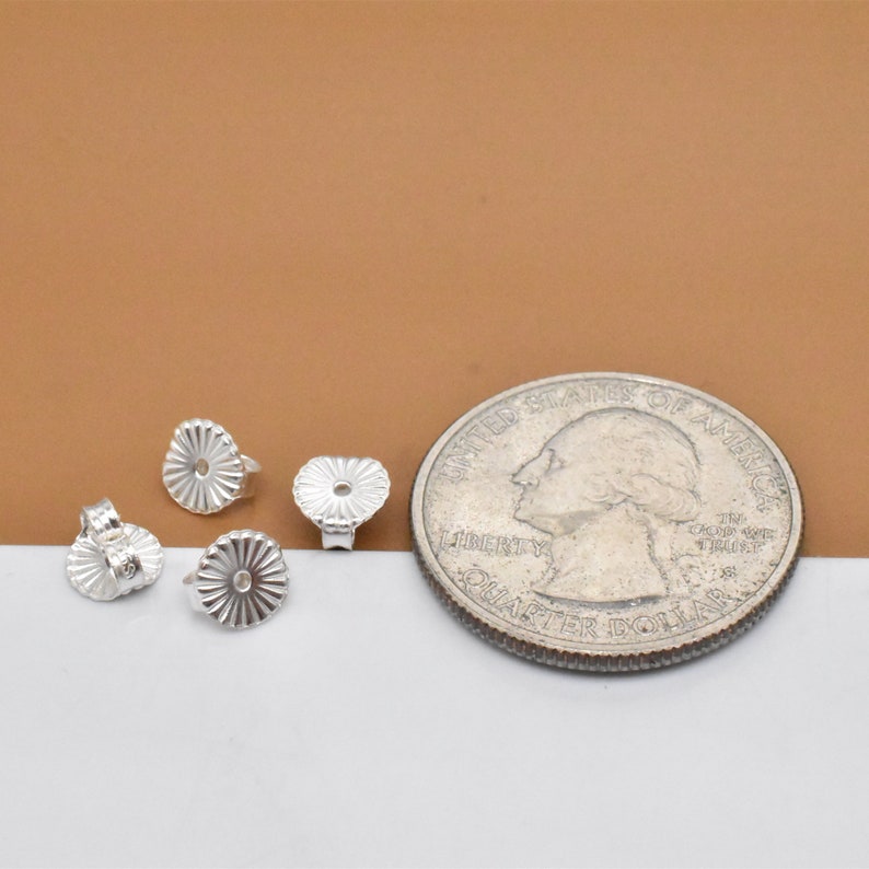 May include: Four silver earring backs, each with a small hole in the center, are arranged on a white surface. A US quarter is included for size comparison.