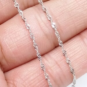 May include: A silver chain necklace with a delicate, twisted design. The chain is shown on a hand, with the ends of the chain hanging down.