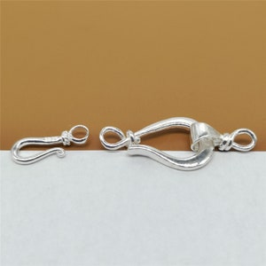 May include: A silver-colored jewelry clasp set, featuring a hook and a curved, ornate clasp. The hook has a small loop at the end, and the larger clasp has a decorative design. The set is designed to connect jewelry pieces.