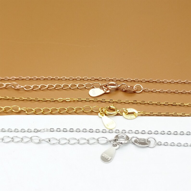 Op de afbeelding: Drie delicate gouden kettingen met karabijnhaken en kleine traanvormige labels. De kettingen liggen op elkaar op een bruin oppervlak.