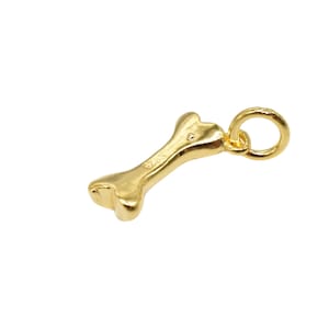 May include: A gold-coloured bone-shaped charm with a small loop for attaching to a necklace or bracelet. The charm has a polished finish and is set against a white background. The charm is likely made of metal and is a dog-themed accessory.