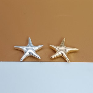Puede incluir: Dos abalorios de estrella de mar de metal, uno plateado y otro dorado. Los abalorios son ambos estrellas de cinco puntas con un centro ligeramente elevado.