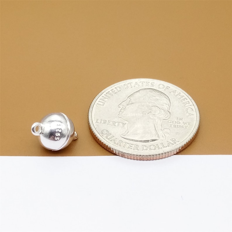 May include: A silver magnetic clasp for jewelry, next to a US quarter for size comparison. The clasp is round and has a smooth surface.